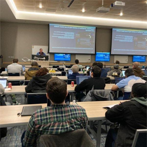 rows of people seated at long tables in classroom, person at front with projected screens behind, about coding and web development